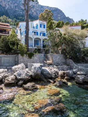 a white house with blue windows next to a body of water at La Veranda Sul Mare in Santa Flavia