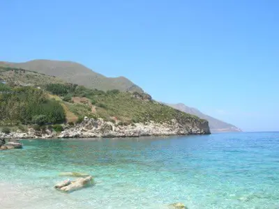a beach with blue water and a mountain in the background at Scopello Beach in Scopello