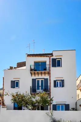 a white building with blue windows and a tree at Santina Suites in Castellammare del Golfo