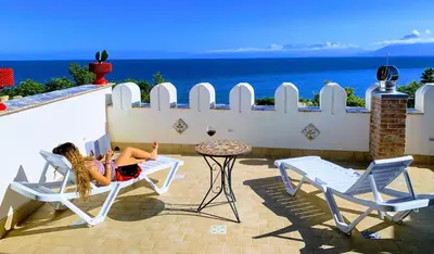 a woman sitting in a chair on a patio overlooking the ocean at Seafront House Gabbiano Azzurro in Santa Flavia