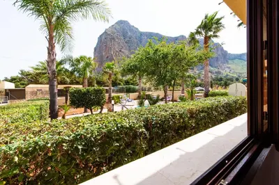 a window view of a resort with palm trees and mountains at Villa Minoa con piscina in San Vito lo Capo